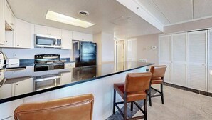 Cozinha moderna com eletrodomésticos de aço inoxidável, uma ilha central com banquetas e vista para um armário branco com portas venezianas.