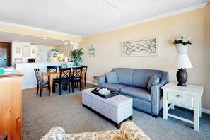 A living room with a grey sofa, a white side table, and a dining area with a wooden table and chairs. - Updated near the beach with balcony, pool, gym & sauna (Ocean City)
