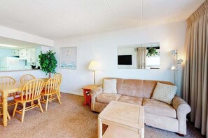 A living room with a sofa, a dining table with chairs, a TV, a lamp, and a plant. - Inviting beach condo with pools, gym, tennis courts & balcony (Ocean City)