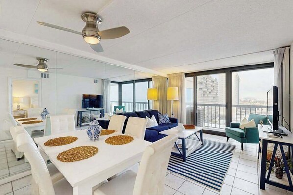 A modern living room with a dining area, a sofa, a TV, and a balcony with a view. - Modern, ocean-view condo with balcony, pools, saunas, tennis & gym (Ocean City)