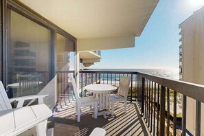 A balcony with a table and chairs, a view of the sea, and a building in the background. - Ocean-view condo with balcony, washer/dryer, pools, saunas & gym (Ocean City)