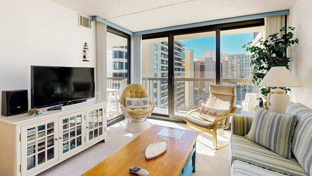 A modern hotel room with a large TV, a sofa, a chair, a balcony with a city view, and a small table. - Ocean-view condo with balcony, washer/dryer, pools, saunas & gym (Ocean City)