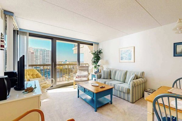 A modern apartment with a balcony, a sofa, a coffee table, a desk with a computer, and a chair. - Ocean-view condo with balcony, washer/dryer, pools, saunas & gym (Ocean City)