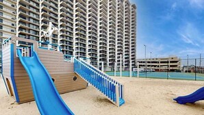 A high-rise building with a playground area featuring a blue slide and a wooden structure. - Beachfront corner condo with balcony, pools, sauna & tennis (Ocean City)