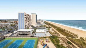 A beachfront hotel with multiple floors, a tennis court, and a beach area with playground equipment. - Beachfront corner condo with balcony, pools, sauna & tennis (Ocean City)