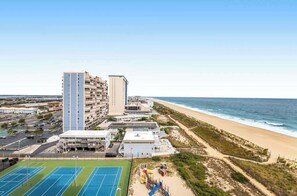 A beachfront hotel with multiple floors, a tennis court, and a beach area with playground equipment. - Beachfront corner condo with balcony, pools, sauna & tennis (Ocean City)