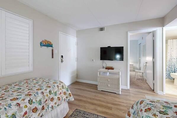 A bedroom with two beds, a television, a white dresser, and a door leading to a bathroom. - Bright, ocean-view condo with balcony, AC, pools, saunas & gym (Ocean City)