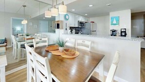 A modern kitchen with white cabinets, a central island, and a dining area with white chairs. - Bright, ocean-view condo with balcony, AC, pools, saunas & gym (Ocean City)