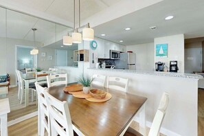 A modern kitchen with white cabinets, a central island, and a dining area with white chairs. - Bright, ocean-view condo with balcony, AC, pools, saunas & gym (Ocean City)