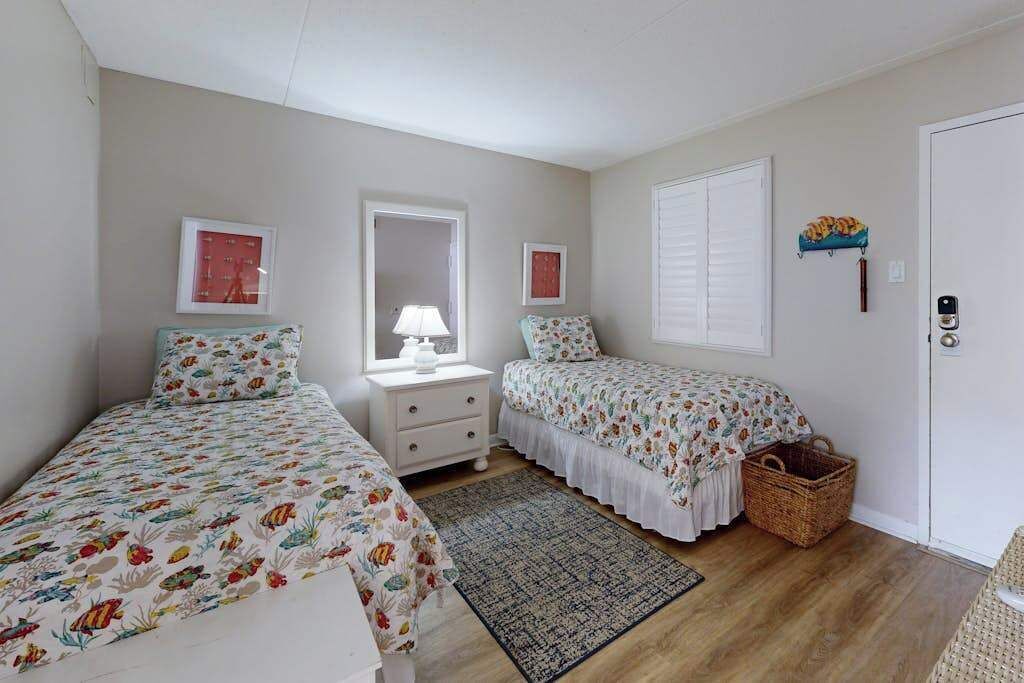 Une chambre avec deux lits, une table de chevet, une lampe, une fenêtre avec des volets, une porte et un panier fixé au mur.