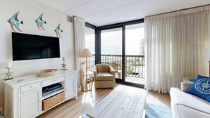 A modern living room with a flat-screen TV, a sofa, a chair, a coffee table, and a large window with curtains. - Bright, ocean-view condo with balcony, AC, pools, saunas & gym (Ocean City)