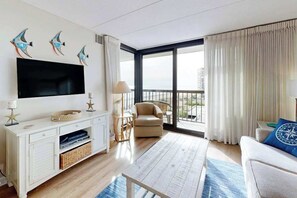 A modern living room with a flat-screen TV, a sofa, a chair, a coffee table, and a large window with curtains. - Bright, ocean-view condo with balcony, AC, pools, saunas & gym (Ocean City)