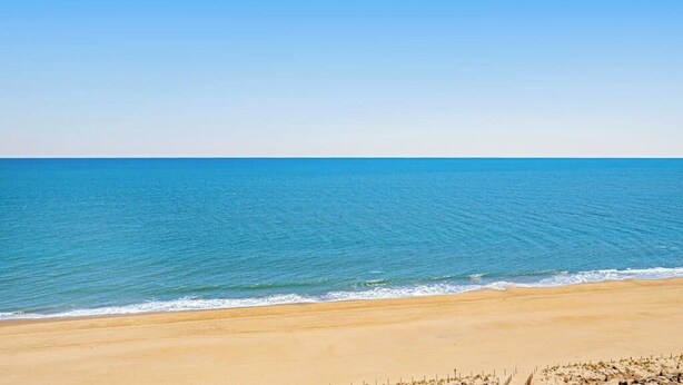 A beach with clear blue water and a sandy shore. - Bright, ocean-view condo with balcony, AC, pools, saunas & gym (Ocean City)