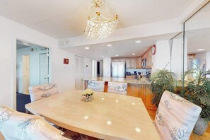 A dining area with a large oval table, chairs with floral upholstery, a chandelier, and a kitchen in the background. - Ocean-view condo with balcony, W/D, pools, saunas, tennis & gym (Ocean City)