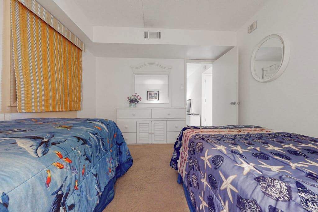 Quarto com duas camas, cómoda, espelho e porta que dá para outro espaço.