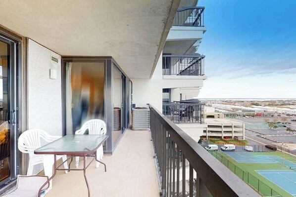 A balcony with a table and chairs, a view of a parking lot, and a building in the background. - Ocean-view condo with balcony, W/D, pools, saunas, tennis & gym (Ocean City)