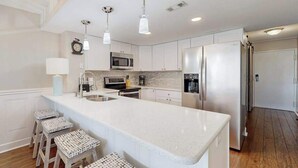 A modern kitchen with white cabinetry, stainless steel appliances, a large island with stools, pendant lighting, and a clock on the wall. - Stylish inviting beach condo with balcony, pools, gym & sauna (Ocean City)