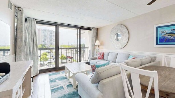 A modern living room with a sofa, coffee table, and dining table. There is a large window with a view of buildings outside. - Stylish inviting beach condo with balcony, pools, gym & sauna (Ocean City)