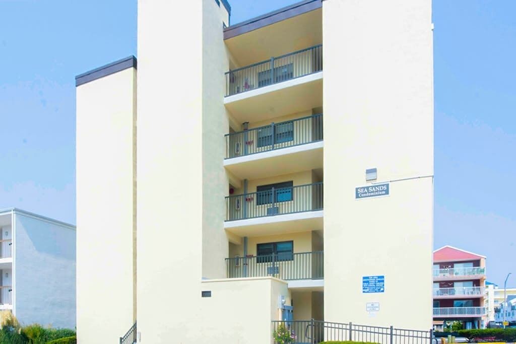 A multi-story building with balconies, a sign indicating 'SEA SANDS Condominiums,' and a clear sky.