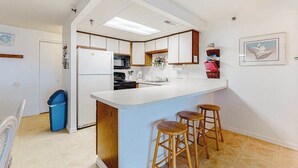 A modern kitchen with a white countertop, wooden stools, and a blue trash bin. - Sandy Square 404 (Ocean City)