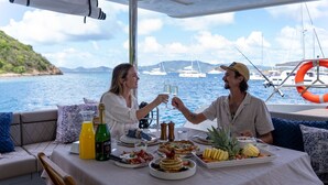 A dining table with food and drinks, a view of the sea, and boats in the background.
