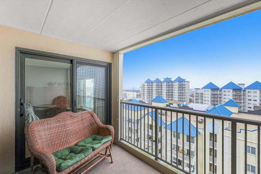 A balcony with a wicker sofa and a glass door, overlooking a residential area with blue-roofed buildings.