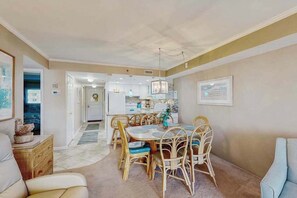 A dining area with a round table and chairs, a kitchen in the background, and a living room with a sofa. - Sandpiper Dunes 302 (Ocean City)