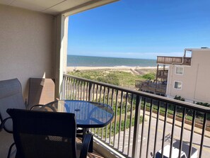 A balcony with a glass table and chairs, overlooking a beach and a building with a sign. - Coastal-Themed Beach Condo w/Great Beach Views, Private Balcony, Shared Pool (Ocean City)