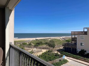 A balcony with a view of the beach, a wooden fence, and a building with a balcony. - Coastal-Themed Beach Condo w/Great Beach Views, Private Balcony, Shared Pool (Ocean City)