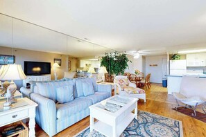 A living room with a sofa, coffee table, and TV, a dining area with chairs, and a kitchen in the background. - Oceanfront, Ground-Floor Condo w/ Patio, Fast WiFi, Central AC & Washer/Dryer (Ocean City)