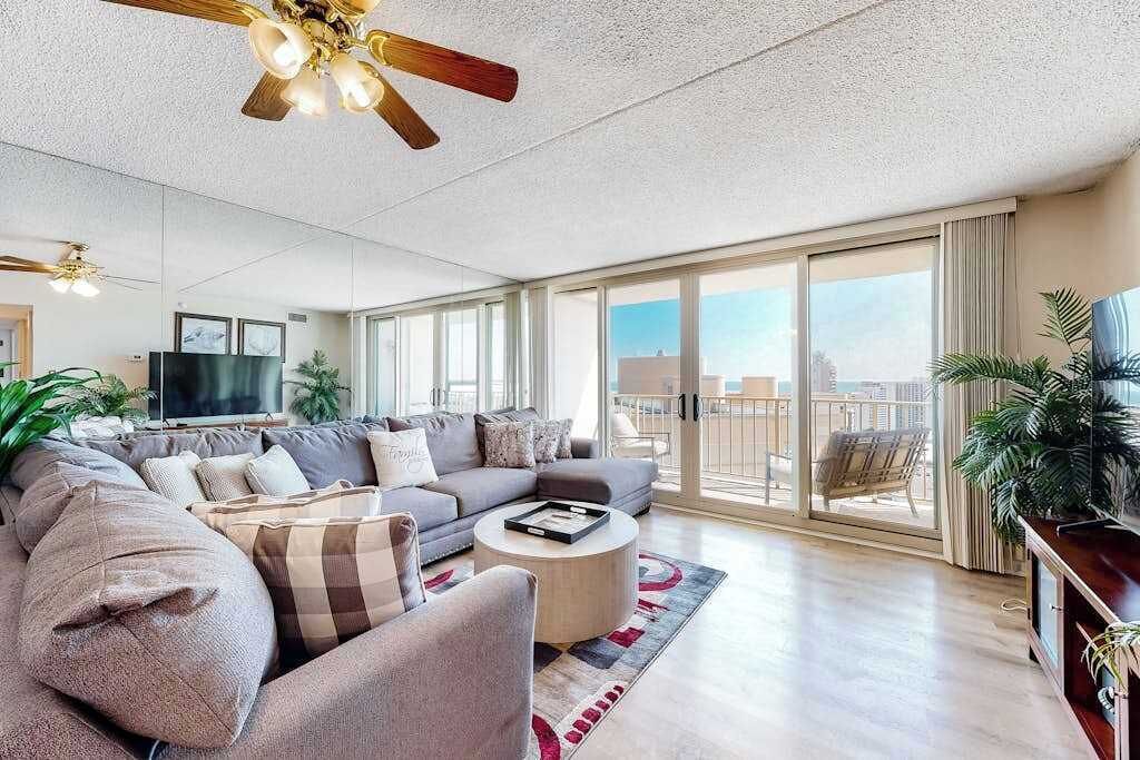 Un salon moderne comprenant un ensemble de canapés, une table basse, un ventilateur de plafond et une grande porte-fenêtre coulissante donnant sur un balcon.