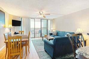 A hotel room with a dining table, chairs, a sofa, a TV, a ceiling fan, and a balcony with a view. - Oceanfront Condo with a Shaded Balcony, AC, WiFi & Shared Outdoor Pool (Ocean City)