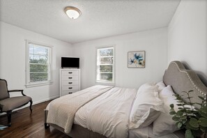 Ein Schlafzimmer mit Bett, Sessel, Kommode, Fernseher und einem Fenster mit Jalousien.