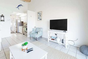 A modern living room with a flat-screen TV, a patterned armchair, a coffee table with a plant, and a kitchen in the background. - Two-Level Bayfront Condo with Amazing Sunsets, Balcony, WiFi & Shared Pool (Ocean City)
