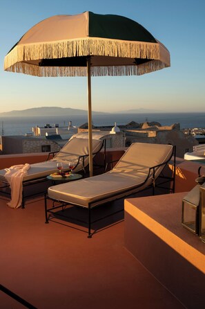 Une terrasse sur le toit avec deux chaises longues, une table avec des boissons et un grand parasol.