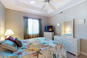 Ein Schlafzimmer mit Bett, Fernseher, Kommode, Deckenventilator und einem Fenster mit Vorhängen.