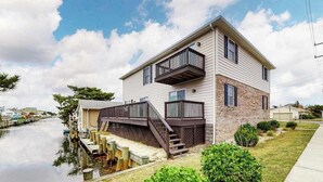Une maison à deux étages avec une jetée qui s’avance dans un plan d’eau, le tout entouré de verdure.