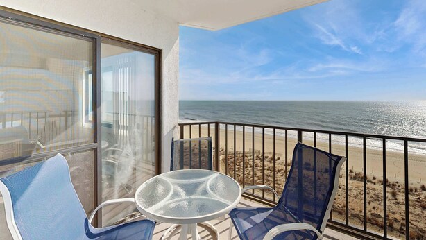 Ein Balkon mit Glastisch und Stühlen, der Blick auf einen Strand mit Zaun und klarem Himmel bietet.