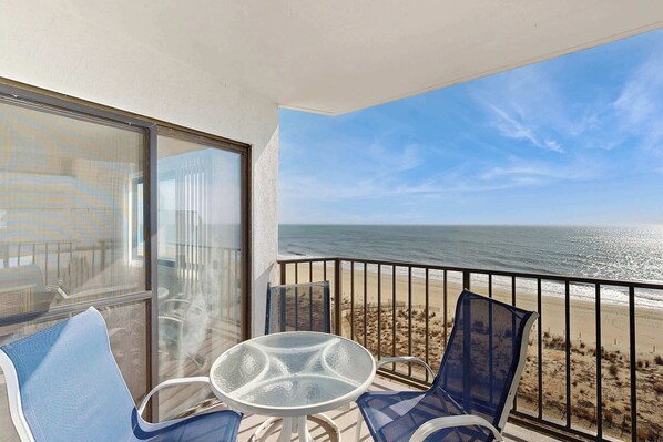A balcony with a glass table and chairs, overlooking a beach with a fence and a clear sky. - Plaza 701 (Ocean City)