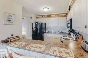 A kitchen with white cabinets, a black refrigerator, a sink, and a countertop with a floral mat. - Convenient Coastal Condo w/Shared Outdoor Pool, Private Washer/Dryer, Free WiFi (Ocean City)