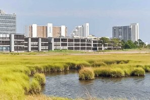 A modern building complex with multiple floors, surrounded by a grassy area and a small water body. - Convenient Coastal Condo w/Shared Outdoor Pool, Private Washer/Dryer, Free WiFi (Ocean City)