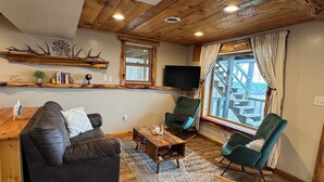 Een gezellige woonkamer met een bank, stoelen, een bijzettafeltje, houten balken in het plafond, een televisie en een raam met uitzicht.