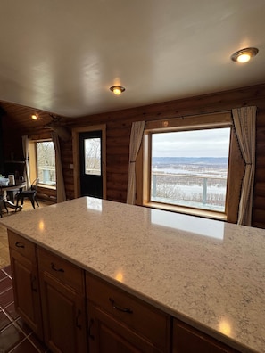 A kitchen with a large island, wooden cabinets, and a view of a snowy landscape through the windows. - Riverview Retreat / Sleeps 6 (De Soto)
