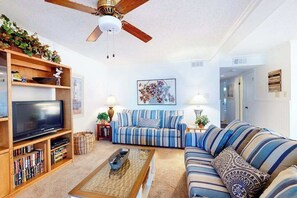 A living room with a television, bookshelf, striped sofa, and ceiling fan. - Our Place At The Beach 106C (Ocean City)