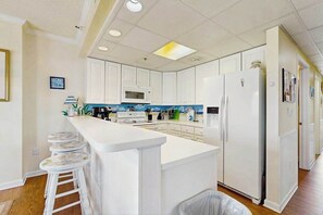 A kitchen with white cabinets, a central island with stools, a refrigerator, and a microwave. - Ocean Block Condo w/ 2 Balconies, AC, Fireplace, Laundry & Shared Pool (Ocean City)