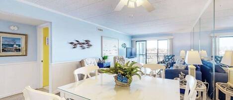 A modern living room with a glass dining table, white chairs, a ceiling fan, and a large window.