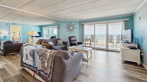 A spacious living room with a large window, a flat-screen TV, and a balcony with chairs. - Airy Beachfront Condo w/Great Views, Private Balcony, W/D, Shared Outdoor Pool (Ocean City)