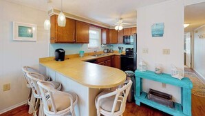A kitchen with wooden cabinets, a central island with bar stools, a ceiling fan, and a teal accent wall. - Top-Floor Condo w/ Shared Deck, Central AC, Washer/Dryer - 1 Block to Beach (Ocean City)