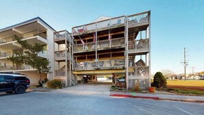 A multi-story building with balconies, a parking area, and a clear sky. - Top-Floor Condo w/ Shared Deck, Central AC, Washer/Dryer - 1 Block to Beach (Ocean City)