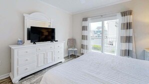 A bedroom with a bed, a television on a dresser, a chair, and a balcony with curtains. - Newly Updated Condo Steps from the Beach w/ Balcony, Shared Rooftop Pool (Ocean City)